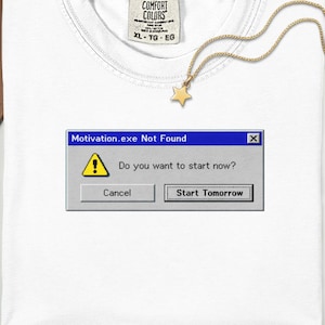 May include: A white t-shirt featuring a graphic of a computer error window with the text "Motivation.exe Not Found" and "Do you want to start now?" with buttons labeled "Cancel" and "Start Tomorrow" alongside a gold star pendant necklace.