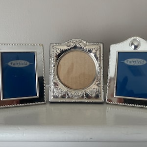 May include: Three silver-toned metal picture frames. Two rectangular frames feature beaded edges and blue inserts with the text Fairfield. The center frame is square with a circular opening, ornate embossed floral details, and a tan insert.