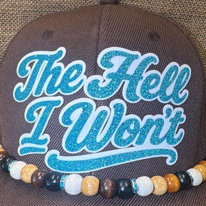 May include: Brown baseball cap with the text "The Hell I Won't" in blue glitter lettering. The cap features a beaded band with various colors including white, brown, black, and blue accents.