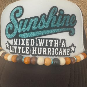 May include: A black and white trucker hat with the word "Sunshine" in teal glitter lettering. The text "Mixed with a Little Hurricane" is below. A beaded necklace with wooden beads and blue rhinestone accents adorns the hat.