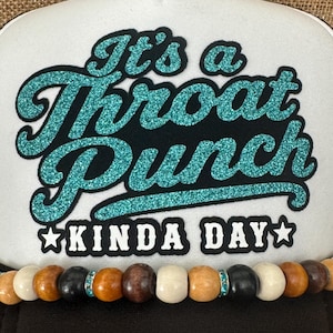 May include: A black and white trucker hat with the text "It's a Throat Punch Kinda Day" in teal glitter lettering. The hat features a beaded band with wooden beads and teal accents.