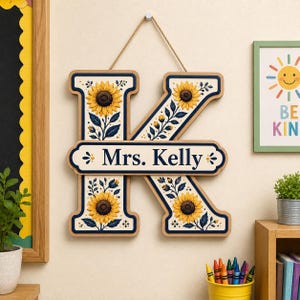 May include: A wooden wall hanging in the shape of the letter K, featuring a cream background with yellow sunflowers and dark blue leaves. A horizontal plaque across the center displays the text Mrs. Kelly in dark blue lettering.