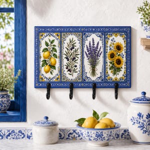 May include: A rectangular wall-mounted hook rack featuring four black metal hooks attached to a ceramic panel. The panel displays four vertical sections with painted illustrations of lemons, olive branches, lavender sprigs, and sunflowers.