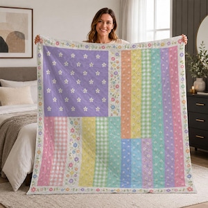 May include: A quilted blanket featuring a patchwork design with a lavender section covered in small white flowers, alongside vertical stripes in pastel yellow, green gingham, light blue, and pink, all bordered by a floral vine pattern.