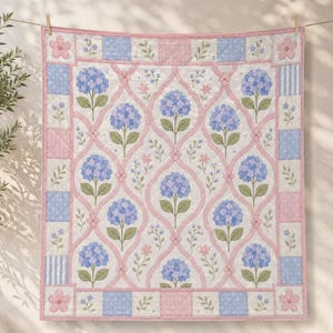 May include: A quilted blanket featuring a repeating pattern of blue hydrangea flowers and small pink blossoms framed by pink lattice lines. The border consists of alternating light blue and pink squares with polka dot and striped fabric patterns.