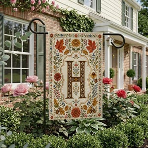 May include: A rectangular garden flag featuring a large, ornate brown letter H in the center, surrounded by a decorative border of colorful autumn leaves, berries, acorns, and flowers on a cream-colored background, hanging on a black metal stand.