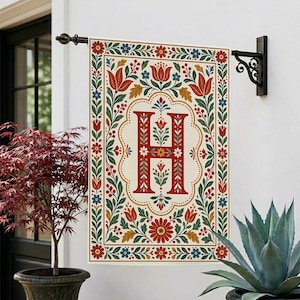 May include: A rectangular garden flag featuring a large, ornate red letter H centered within a cream-colored field. The design is framed by a decorative border of red, blue, and green floral and leaf motifs on a white background.