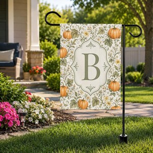 May include: A rectangular garden flag featuring a central ornate frame enclosing the letter B in a serif font. The background displays a botanical pattern of orange pumpkins, green leaves, and small yellow flowers on a light cream-colored fabric.