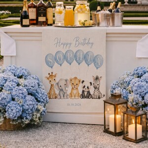 May include: A rectangular beige fabric banner featuring the text Happy Birthday and the name SOPHIA printed in blue letters inside six light blue balloons. Below, illustrations of a giraffe, elephant, lion, panda, and zebra are displayed with the date 10.10.2026 printed at the bottom.