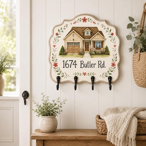 May include: A scalloped-edge wall-mounted sign featuring a painted illustration of a house, the text 1674 Butler Rd., and a floral vine border. The sign includes four black metal hooks at the bottom for hanging items, with a cream-colored background.