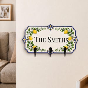 May include: A rectangular wall-mounted hook rack with a cream-colored ceramic plaque featuring a blue border, green lemon tree branches with yellow lemons, and the text THE SMITHS in black. It has three black metal hooks attached.