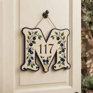 May include: A cream-colored, letter M-shaped wall plaque featuring the number 117 in black. The letter is decorated with painted green leaves, white flowers, and dark blue blueberries. It hangs from a dark brown hook by a thin rope.