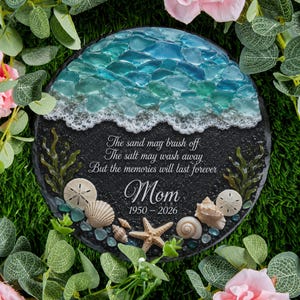 Pode incluir: Uma placa memorial preta redonda com uma seção superior de vidro do mar azul e turquesa imitando ondas com espuma branca. O texto central diz: The sand may brush off, The salt may wash away, But the memories will last forever, Mom, 1950-2026. Decorada com conchas e uma estrela-do-mar.