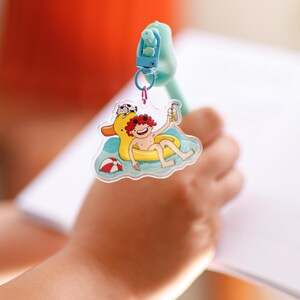 Peut inclure: Un porte-clés en acrylique transparent présentant une illustration de dessin animé d'une personne aux cheveux roux flottant dans une bouée canard jaune sur l'eau bleue, tenant une boisson, avec un petit ballon de plage et un chat à proximité.