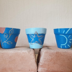 Puede incluir: Tres macetas pequeñas de terracota pintadas en tonos azules. La maceta izquierda presenta un diseño de enredadera dorada con círculos rosas. La central muestra una estrella de mar dorada sobre fondo azul claro. La derecha tiene un sol azul claro.