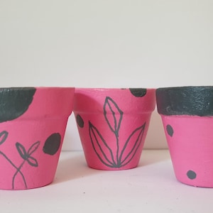 Könnte beinhalten: Fünf kleine, konische Blumentöpfe, die in leuchtendem Pink mit dunkelgrauen Akzenten bemalt sind. Jeder Topf weist ein einzigartiges handgemaltes Design auf, darunter dunkelgraue Punkte, feine Blattumrisse und kurze horizontale Pinselstriche.