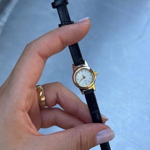 May include: A gold-tone analogue wristwatch featuring a small round white face with gold markers and hands. The watch has a slim black textured leather strap with a gold-tone buckle closure. The dial displays the word Quartz at the bottom.