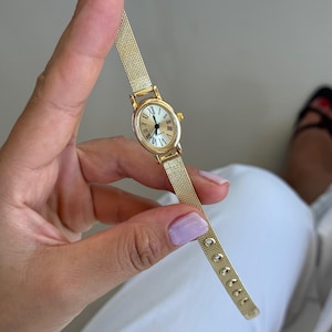 Può includere: Un orologio da polso color oro con cassa ovale, quadrante color crema, numeri romani neri e lancette nere. L'orologio include un cinturino in maglia metallica color oro fine con un sistema di chiusura a fibbia regolabile.