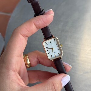 Könnte beinhalten: Eine rechteckige Armbanduhr mit weißem Zifferblatt, schwarzen Stundenmarkierungen und dem in Schwarz gedruckten Wort QUARTZ. Die Uhr hat ein goldfarbenes Metallgehäuse und ein dunkelbraunes Lederarmband mit goldfarbener Schließe und Beschlägen.