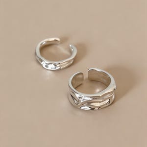 Può includere: Due anelli color argento con design aperto. Ogni anello presenta una superficie lucida e riflettente con rientranze organiche irregolari e contorni fluidi e ondulati che creano un aspetto strutturato sulle fasce metalliche.
