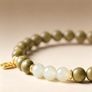 Könnte beinhalten: Ein Perlenarmband mit glatten, olivgrünen, kugelförmigen Perlen und drei zentralen, durchscheinenden weißen Perlen. Das Design umfasst kleine, polierte goldfarbene Metallabstandshalter zwischen den Perlen und einen kleinen, flachen goldfarbenen Anhänger.
