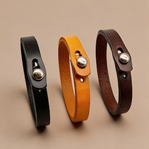 Può includere: Tre braccialetti in pelle nei colori nero, ocra e marrone scuro. Ogni braccialetto presenta un design a fascia piatta e liscia con una chiusura a bottone a pressione in metallo argentato. Le fasce sono disposte verticalmente per mostrare i colori.