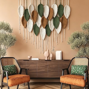 Può includere: Un arazzo in macramè con molteplici elementi in fibra a forma di foglia nei colori crema, verde bosco e marrone terracotta, sospesi a un bastone di legno orizzontale con una corda bianca per il fissaggio, creando una disposizione a strati.
