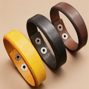Könnte beinhalten: Drei Lederarmbänder in Gelb, Schwarz und Braun. Jedes Armband hat eine glatte, matte Oberfläche und wird mit zwei silberfarbenen Metalldruckknöpfen auf der Innenseite befestigt, was einen verstellbaren Verschluss und ein schlichtes Design ermöglicht.