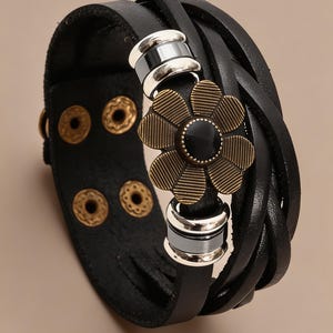 Op de afbeelding: Een zwarte armband met meerdere strengen en een centrale bronzen bloembedel met een zwarte steen in het midden. Het ontwerp bevat zilverkleurige metalen tussenstukjes en een brede zwarte band die wordt vastgezet met twee paar messingkleurige drukknopen.