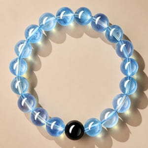 Può includere: Un bracciale circolare composto da perline sferiche traslucide di colore azzurro disposte in un cerchio continuo, con una singola perla sferica nera, solida e opaca, posizionata al centro della parte inferiore.