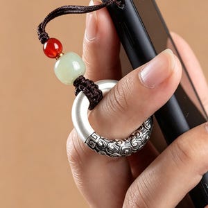 May include: A phone charm featuring a dark brown braided cord attached to a black smartphone. The charm includes a smooth red bead, a pale green bead, and a silver-toned ring with an intricate embossed cloud pattern on its lower half.