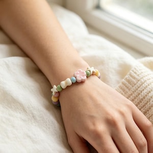 Può includere: Un braccialetto di perline con una perla centrale a forma di fiore rosa chiaro, intervallata da perline rotonde più piccole in tonalità pastello blu, verde, giallo e viola, insieme a perline bianche a forma di stella e fiocco su un cordino elastico.