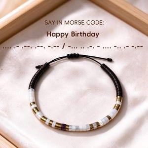 Può includere: Un braccialetto di perline con cordino nero e nodo regolabile, decorato con una sequenza di perline marrone scuro, bianche, oro metallizzato e una singola perlina viola. Il testo recita: SAY IN MORSE CODE: Happy Birthday, seguito dal codice Morse.