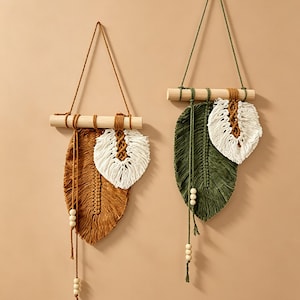Puede incluir: Dos tapices de macramé con formas de hojas suspendidas de varas de madera. La pieza de la izquierda es de color amarillo mostaza con un acento blanco, mientras que la derecha es verde oliva con una hoja blanca. Cuentas de madera en cordones.