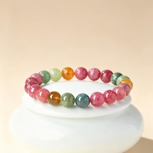 Può includere: Un braccialetto elastico composto da perline di pietre preziose lisce, rotonde e levigate in una varietà di colori, tra cui sfumature di rosa, rosso intenso, verde bosco, verde oliva e giallo ambra, disposte in un motivo circolare.