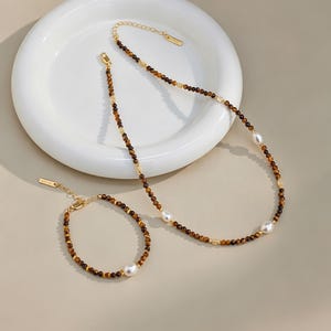 Può includere: Un set composto da collana e bracciale con perline in occhio di tigre marrone, piccoli distanziatori dorati e perle bianche. Entrambi i pezzi includono una catenella di estensione dorata con una piccola targhetta metallica rettangolare sul fermaglio.