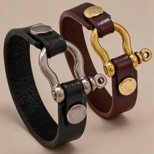 May include: Two leather bracelets, one black and one dark brown, each featuring a metal shackle clasp. The black bracelet has a silver-toned shackle and matching stud, while the brown bracelet has a brass-toned shackle and matching stud.