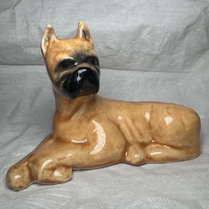 May include: A ceramic figurine of a Boxer dog in a lying position. The dog has a light tan coat with a glossy finish, featuring a distinct black muzzle and dark shading around the eyes and ears. The sculpture captures the alert posture of the breed.