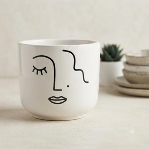 Puede incluir: Una maceta de cerámica blanca con un dibujo de líneas negras que representa un rostro. El diseño incluye un ojo cerrado con pestañas, una línea de nariz, un pequeño punto y labios. La maceta tiene una forma cilíndrica lisa y abierta.