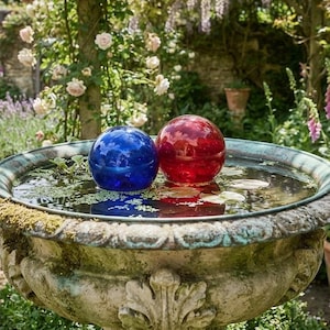 May include: A weathered stone birdbath with a wide, circular basin filled with water. Two smooth, spherical glass gazing balls, one deep blue and one vibrant red, float on the surface of the water. The basin features a decorative, carved rim.
