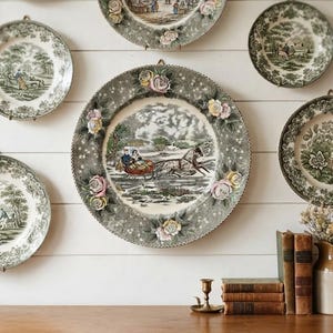 May include: A collection of round ceramic decorative plates featuring green transferware landscape scenes and applied floral accents, displayed on a white wall above a wooden surface with a stack of three books and a ceramic pitcher.