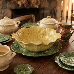 May include: A collection of ceramic tableware featuring cabbage leaf designs. The set includes a large yellow cabbage-shaped bowl, several green cabbage-leaf plates, cream-colored lidded tureens, and a multi-colored pitcher with leaf motifs.