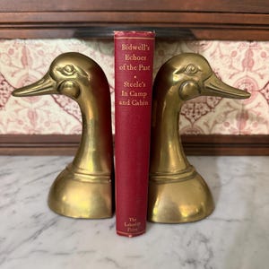 May include: Two polished brass duck head bookends flanking a red hardcover book titled Bidwell's Echoes of the Past, Steele's In Camp and Cabin, published by The Lakeside Press. The bookends feature detailed eyes and beaks facing outward.