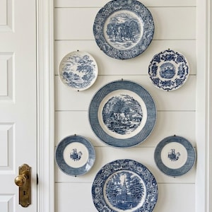 May include: A collection of six blue and white ceramic plates arranged on a wall. The plates feature various detailed landscape and architectural scenes, including trees, buildings, and figures, with intricate floral borders on the edges.