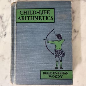 May include: A vintage hardcover book titled Child-Life Arithmetics by Breed, Overman, and Woody. The blue cloth cover features a green rectangular graphic with black text and an illustration of a figure holding a bow and arrow in green and black.
