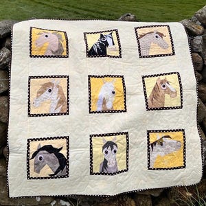 Op de afbeelding: Een crèmekleurige quilt met negen vierkante panelen, elk met een ander paardenhoofdontwerp. De panelen hebben een gele achtergrond en zijn omlijst met een zwart-witte stippenrand. De rand van de quilt is ook afgewerkt met dezelfde stippenstof.