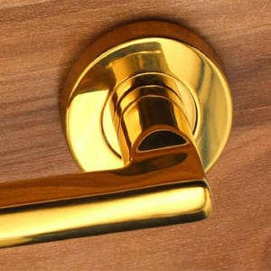May include: A polished gold-tone metal door handle featuring a circular mounting plate and a horizontal lever. The hardware has a smooth, reflective surface and is attached to a flat, light brown wood-grain surface with visible horizontal texture.