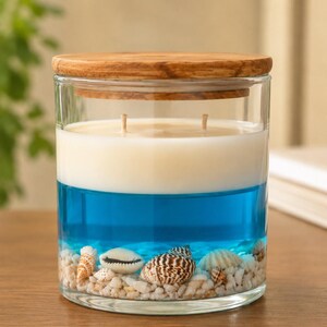 Peut inclure: Un pot à bougie en verre transparent avec un couvercle en bois, doté de deux mèches, une couche de cire blanche, une couche de gel bleu et une base remplie de petits galets blancs et de divers coquillages, dont un cauri et un coquillage spiralé.