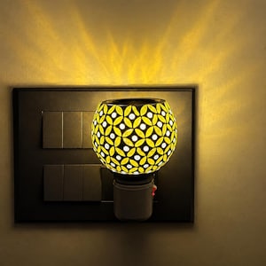 Peut inclure: Un diffuseur mural à brancher doté d'un abat-jour sphérique en verre avec un motif floral en mosaïque jaune et blanc. L'appareil est branché sur une plaque murale rectangulaire sombre avec plusieurs interrupteurs, diffusant une lumière chaude.