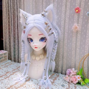 May include: A pale lavender wig with cat ears styled on a mannequin head. The hair features two thick braids, silver star and bow hair clips, and a white lace collar necklace. The mannequin has large blue eyes and a pale complexion with facial markings.
